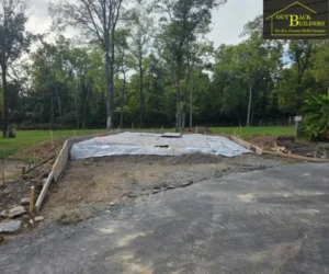 The preparation site for a concrete project, featuring wooden forms and a protective plastic liner laid over a gravel base near an existing asphalt path.