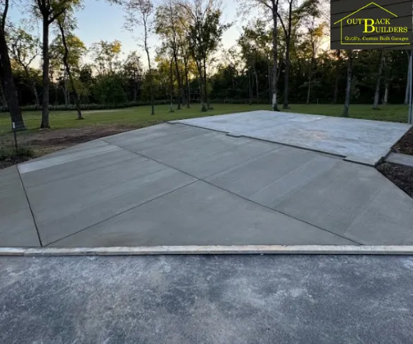 A freshly poured and smoothed concrete driveway extension and garage pad in a grassy area bordered by trees.
