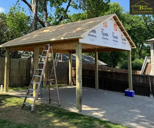 carport under construction with exposed roof framing, weather-barrier wrap on the gable end, and ladders on a concrete slab