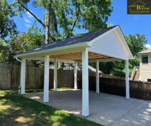 Completed open-air wooden carport featuring white support posts, a dark shingled gabled roof, and a concrete driveway